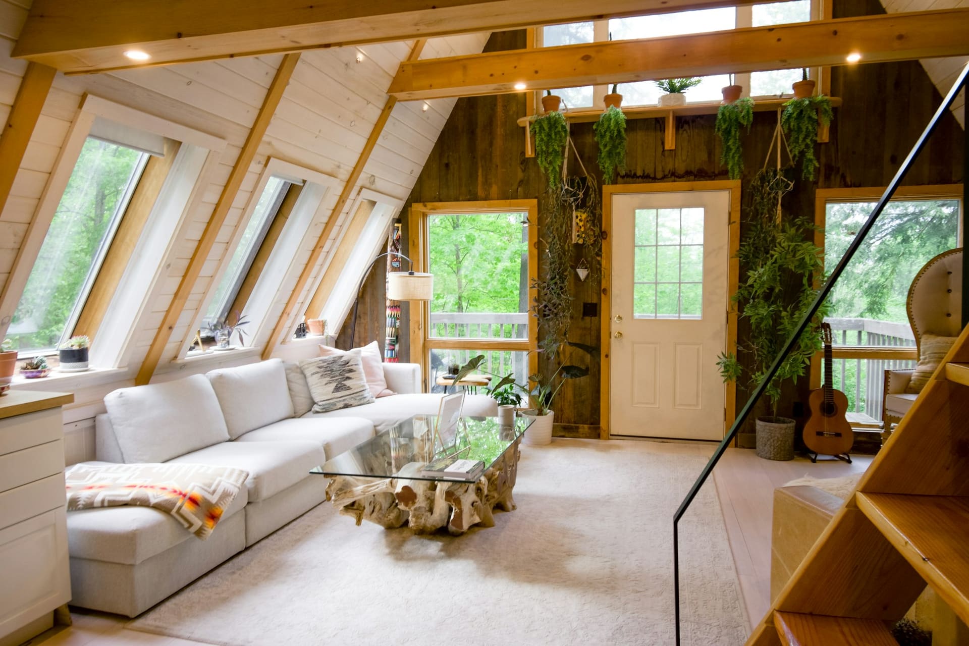 An A-frame cabin living room with a long white sofa, a glass coffee table on a driftwood base, timber walls and hanging plants.