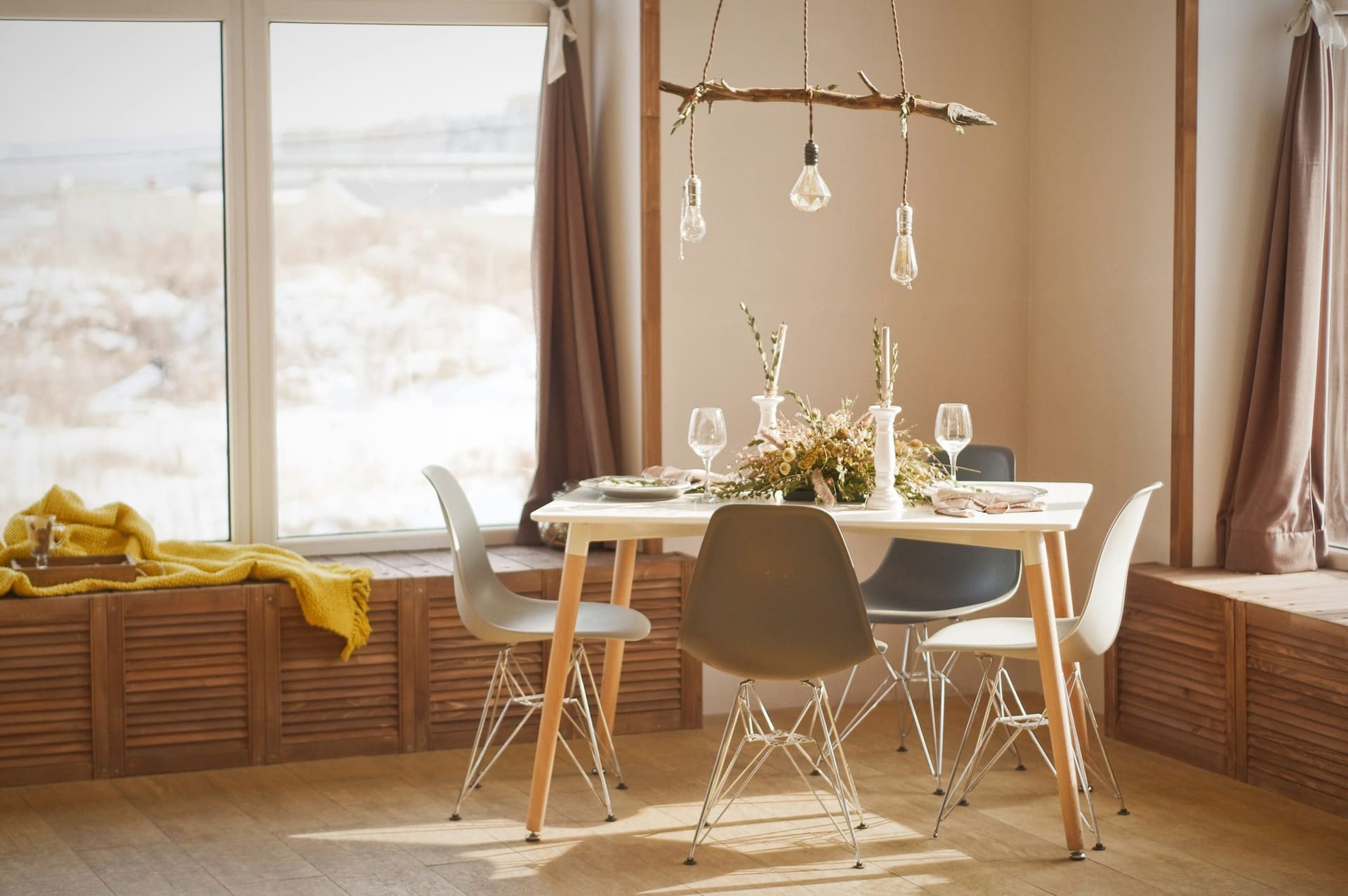 A dining table with white moulded chairs in front of a window seat, a branch pendant hung with bare bulbs above.