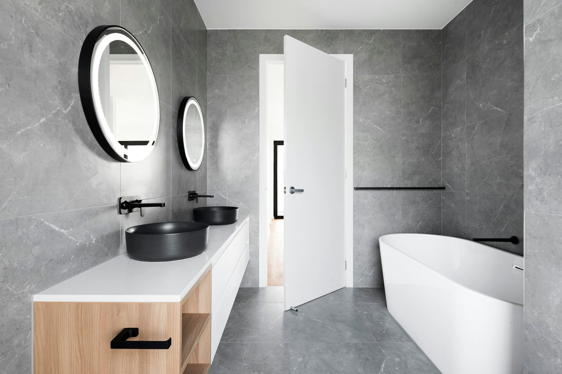 A grey stone-effect bathroom with a freestanding bath, round backlit mirrors and an oak-fronted vanity with a black basin.