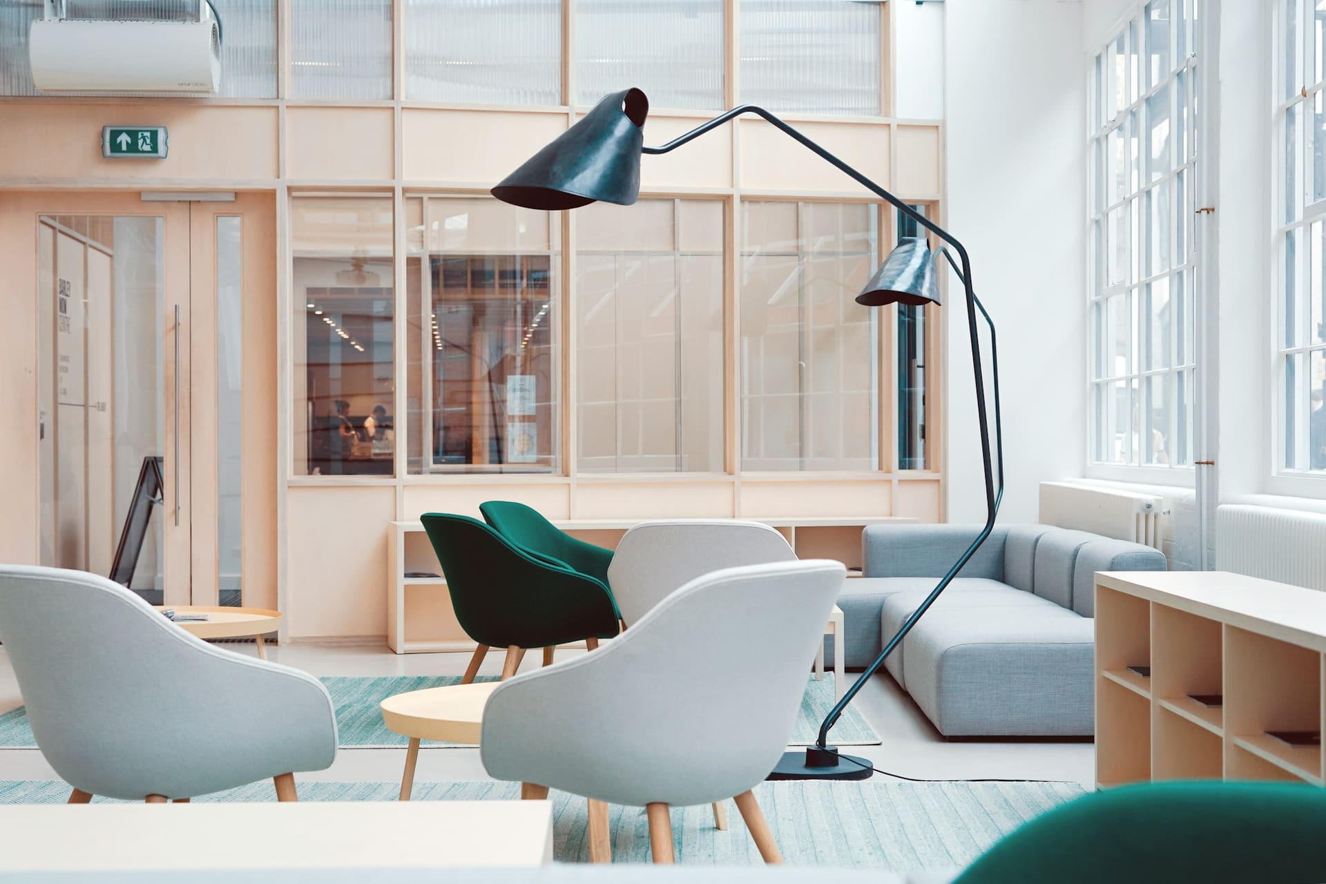 A bright workspace with white lounge chairs around a low table, a black task lamp and shelving below steel-framed windows.