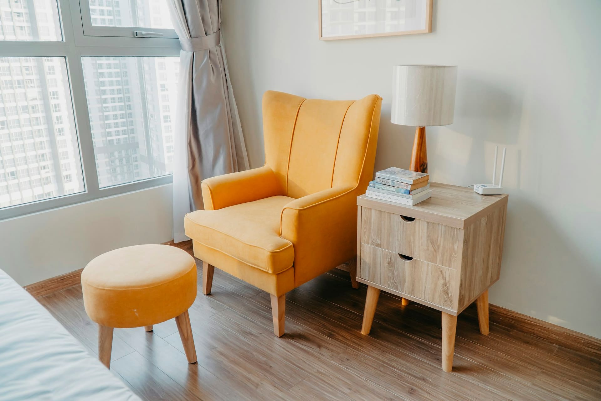 A mustard wingback armchair and matching footstool beside an oak bedside table and a white ceramic lamp.