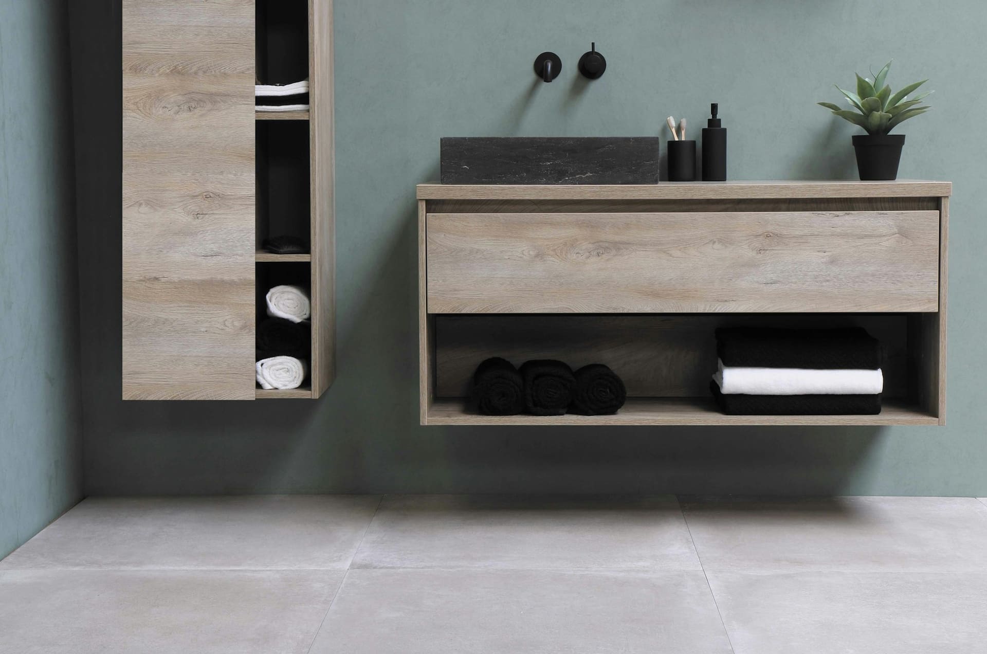 A wall-hung oak vanity with an open shelf of rolled towels against a sage-green wall and a concrete floor.