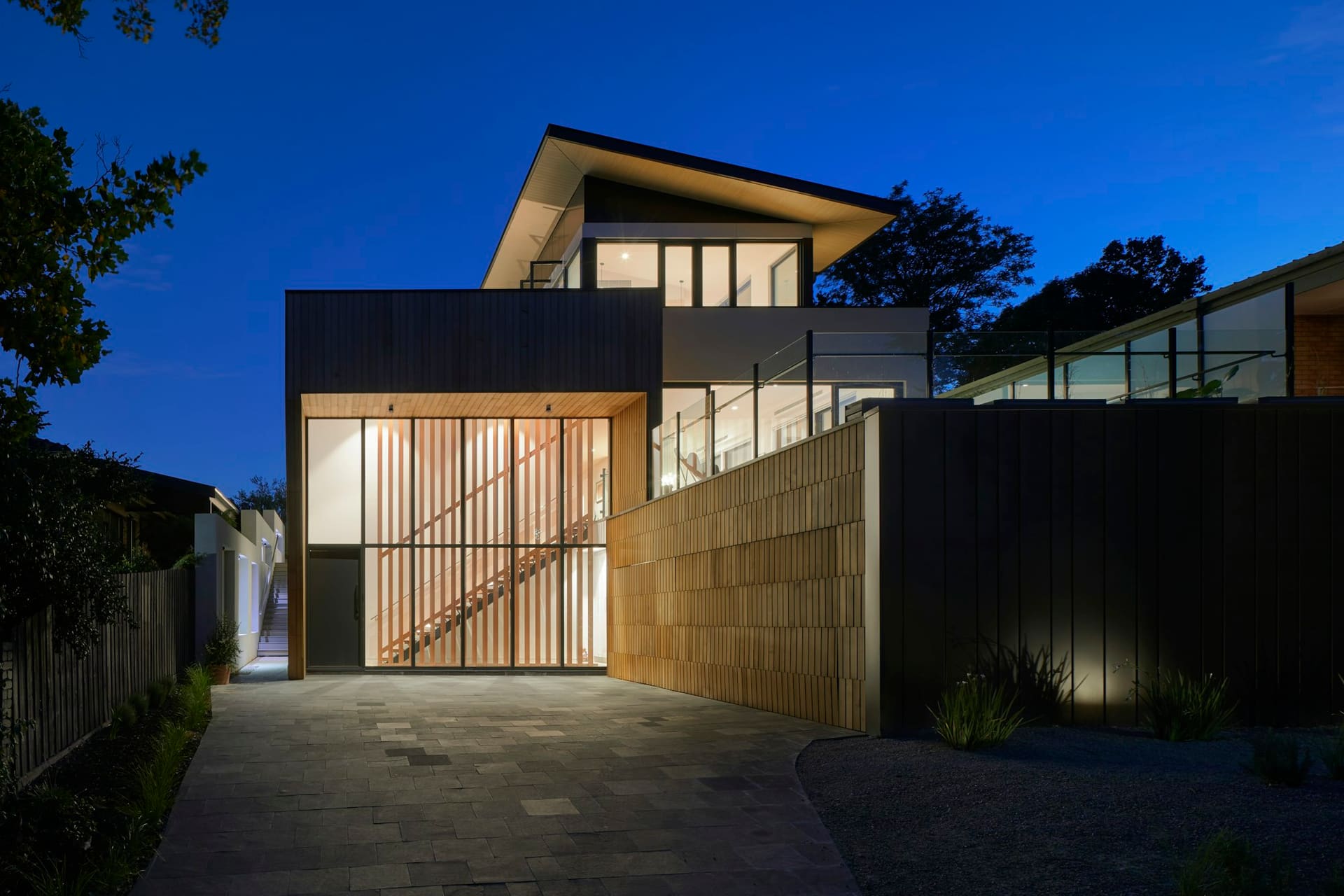 A contemporary house at dusk with a timber-clad stair tower lit from inside and a rendered garden wall.